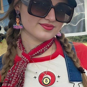 May include: A person wearing a black cap, large sunglasses, and earrings. They are wearing a red and white baseball tee, denim overalls, and a red bandana. A necklace with an 8-ball pendant is visible.