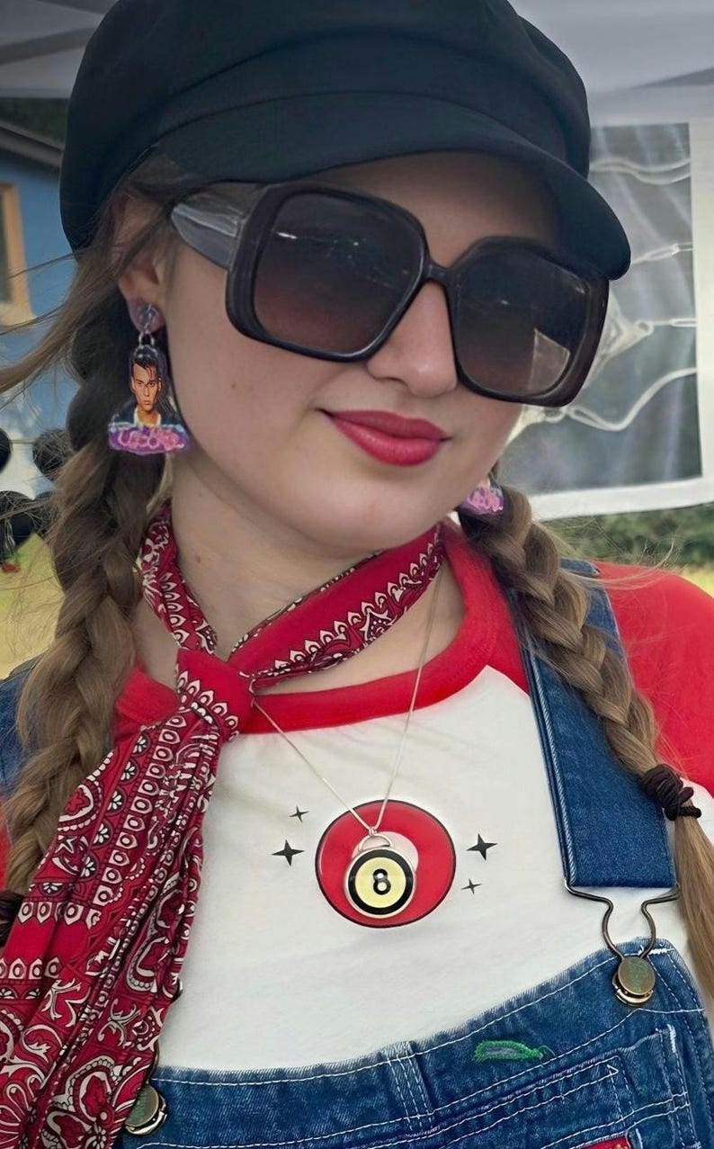 May include: A person wearing a black cap, large sunglasses, and earrings. They are wearing a red and white baseball tee, denim overalls, and a red bandana. A necklace with an 8-ball pendant is visible.