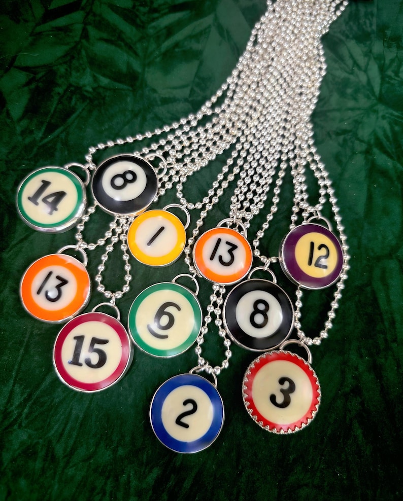 May include: A selection of silver ball chain necklaces, each with a pendant resembling a billiard ball. The pendants show numbers 1, 2, 3, 6, 8, 12, 13, 14, and 15, with different coloured borders.