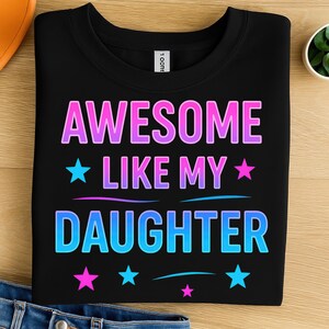 May include: Black t-shirt with the text "AWESOME LIKE MY DAUGHTER" in a gradient of pink and blue. The design includes stars and curved lines. The shirt is folded on a wooden surface.