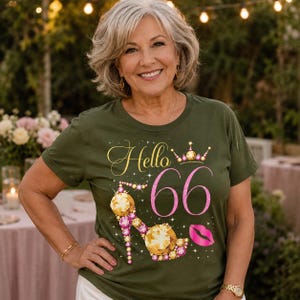 Puede incluir: Una camiseta de manga corta color verde oliva con el texto Hello 66 en letra dorada y rosa. El gráfico incluye un zapato de tacón decorado con gemas doradas y rosas, una pequeña corona dorada, un icono de labios rosas y estrellas blancas.