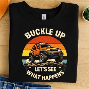 Könnte beinhalten: Ein schwarzes T-Shirt mit Rundhalsausschnitt und einer Grafik eines orangefarbenen Geländewagens auf felsigem Untergrund. Das Design zeigt einen Sonnenuntergang mit horizontalen Streifen und den Text BUCKLE UP LET'S SEE WHAT HAPPENS in Cremefarbe.
