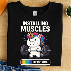 May include: Black sweatshirt featuring the text "INSTALLING MUSCLES" and a cartoon unicorn lifting weights. The unicorn has a rainbow mane and a yellow horn. Below the unicorn is the text "PLEASE WAIT..." in a rainbow bar.