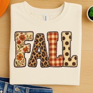 May include: A cream-colored crew neck t-shirt featuring the word FALL in large block letters. Each letter has a distinct pattern: the F is floral, the A is leopard print, the L is orange gingham, and the L is cream with brown polka dots.
