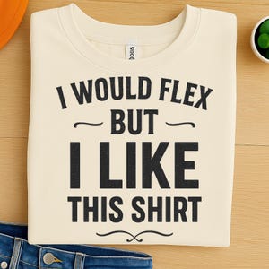 May include: Cream-colored sweatshirt with the text "I WOULD FLEX BUT I LIKE THIS SHIRT" in black block letters. The shirt has a crew neck and is folded neatly. A small white tag is visible near the neckline.