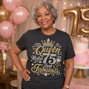 Puede incluir: Una camiseta de manga corta gris oscuro con un diseño gráfico de pedrería dorada y plateada. El texto dice This Queen Makes 75 Look Fabulous, acompañado de una ilustración de una corona y detalles de diamantes en el pecho.
