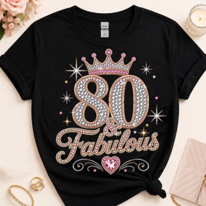 Op de afbeelding: Een zwart T-shirt met korte mouwen en een grafisch ontwerp met de tekst 80 & Fabulous. Het ontwerp bevat een met juwelen versierde kroon, fonkelende sterren en een roze hart, alles uitgevoerd in een diamantachtige textuur met gouden omlijningen.