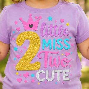 May include: A light purple short-sleeved t-shirt featuring a graphic print with a gold glitter number 2, a pink crown, and the text little MISS Two CUTE in purple, blue, and silver glitter. The design includes small pink, blue, and yellow stars and hearts.