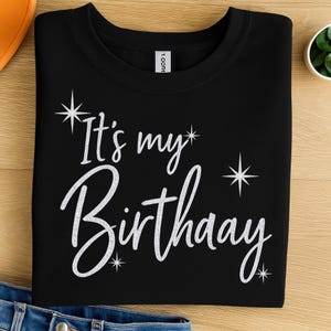 May include: A black crew neck t-shirt featuring the white text "It's my Birthaay" in a cursive script font. The design is accented with several four-pointed star shapes scattered around the lettering, creating a festive and celebratory appearance.