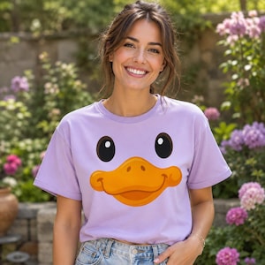 Puede incluir: Una camiseta de manga corta en color violeta claro con un gráfico grande y centrado de una cara de pato de dibujos animados. El diseño incluye dos ojos negros circulares situados sobre un pico de pato naranja brillante y sonriente.