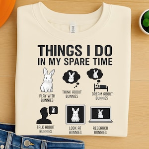 May include: Cream-coloured sweatshirt featuring the text "THINGS I DO IN MY SPARE TIME" and bunny illustrations. The design includes a bunny, thought bubbles, a bed, a person talking, a computer screen, and the phrases "PLAY WITH BUNNIES", "THINK ABOUT BUNNIES", "DREAM ABOUT BUNNIES", "TALK ABOUT BUNNIES", "LOOK AT BUNNIES", and "RESEARCH BUNNIES".