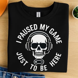 May include: Black crew neck t-shirt with a white graphic. The graphic features a skull wearing headphones and sunglasses, with the text "I PAUSED MY GAME JUST TO BE HERE". A video game controller is also in the graphic.