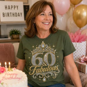 Könnte beinhalten: Ein olivgrünes Kurzarm-T-Shirt mit einem Grafikdruck, der ein juwelenbesetztes Diadem über der Zahl 60 und dem Text & Fabulous zeigt. Das Design ist in Silber- und Goldtönen gehalten und mit funkelnden Sternakzenten versehen.