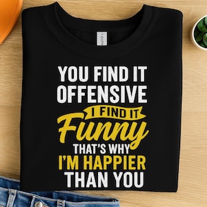 Op de afbeelding: Zwart T-shirt met de tekst "YOU FIND IT OFFENSIVE I FIND IT FUNNY THAT'S WHY I'M HAPPIER THAN YOU" in witte en gele letters. Het shirt is opgevouwen op een houten oppervlak.