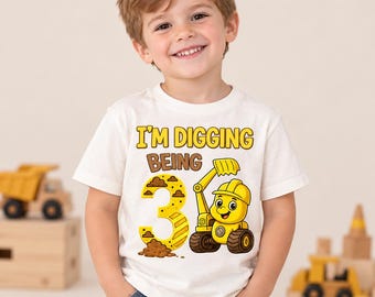 Puede incluir: Una camiseta blanca de manga corta con un gráfico de una excavadora de construcción amarilla y el número tres. El texto dice I'M DIGGING BEING 3 en letras amarillas y marrones. El diseño incluye montones de tierra y un casco de construcción.