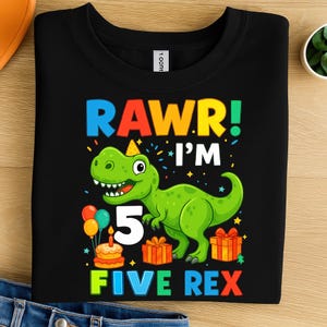 Puede incluir: Camiseta negra con un dinosaurio verde, el número 5 y el texto "RAWR! I'M 5 FIVE REX". El diseño incluye globos, una tarta de cumpleaños y cajas de regalo. La camiseta es de material suave.