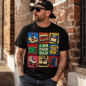 Może przedstawiać: Czarny t-shirt z siatką dziewięciu kolorowych ikon retro, w tym joysticka, kasety magnetofonowej, dyskietki, telewizora, telefonu z tarczą, wrotki, aparatu fotograficznego i odtwarzacza muzyki z napisem I AM THIS OLD pośrodku.
