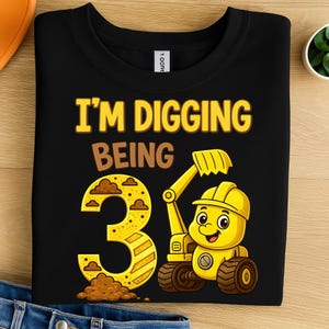 以下が含まれることがあります： 「I'M DIGGING BEING 3」の文字が入った黒いクルーネックTシャツ。数字の3は黄色と茶色のアクセントで、黄色の漫画の掘削機が数字の右側にあります。