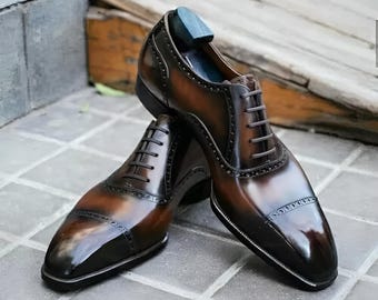 May include: A pair of brown leather dress shoes with a glossy finish. The shoes have a classic Oxford design with a cap toe and lace-up closure. The leather has a gradient effect, transitioning from a lighter brown to a darker shade.