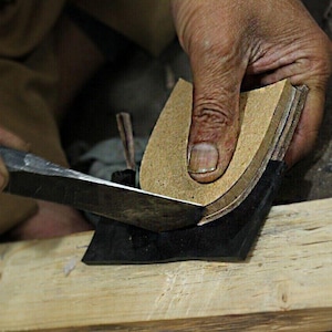 May include: A close-up shot of a shoemaker using a knife to trim the edges of a leather shoe sole. The sole is held in place on a wooden block. The shoemaker's hands are visible, demonstrating the craft of shoe making.