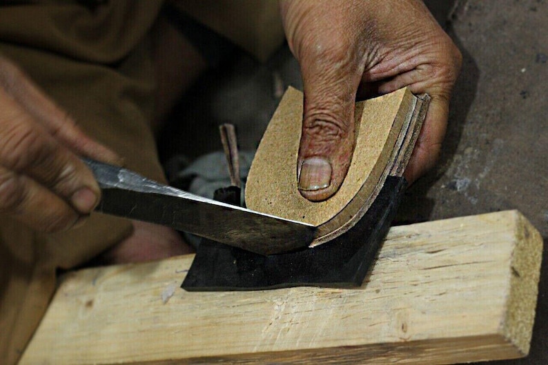 May include: A close-up shot of a shoemaker using a knife to trim the edges of a leather shoe sole. The sole is held in place on a wooden block. The shoemaker's hands are visible, demonstrating the craft of shoe making.