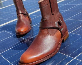May include: A pair of brown leather ankle boots with a harness strap and silver-tone hardware. The boots have a pointed toe and a low heel. The leather has a polished finish and a slight color variation.