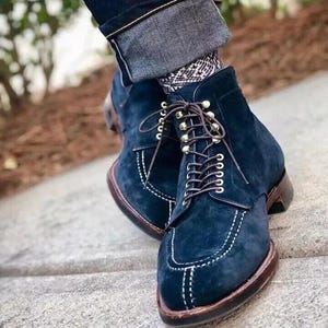 May include: A pair of navy blue suede ankle boots with white stitching and brown leather soles. The boots have brown laces and metal eyelets. The boots are worn with patterned socks and dark denim jeans.