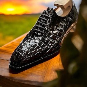 May include: A black, textured leather dress shoe with a lace-up design. The shoe has a glossy finish and is displayed on a wooden shoe tree. The background features a blurred sunset over a green field.