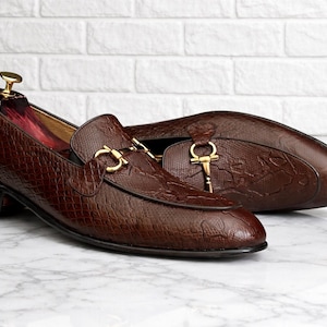 May include: A pair of brown leather loafers with a textured pattern. The shoes feature gold-colored hardware and a classic design. One shoe has a shoe tree inside. The loafers are displayed on a marble surface.