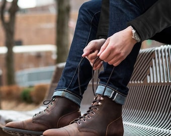 May include: Brown leather boots with cap toes and black laces. The boots are being laced up by a person wearing dark blue jeans. The jeans are cuffed at the ankles, revealing a white and blue striped lining. A watch is visible on the person's wrist.