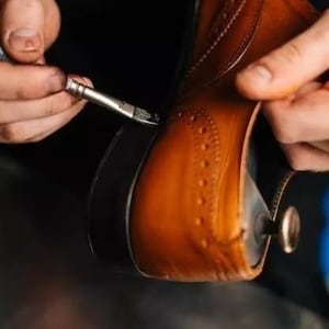 May include: Close-up of a brown leather shoe being worked on with a small brush. The shoe has decorative perforations and a dark sole. The shoemaker is applying a dark substance to the shoe's edge, highlighting the craftsmanship.