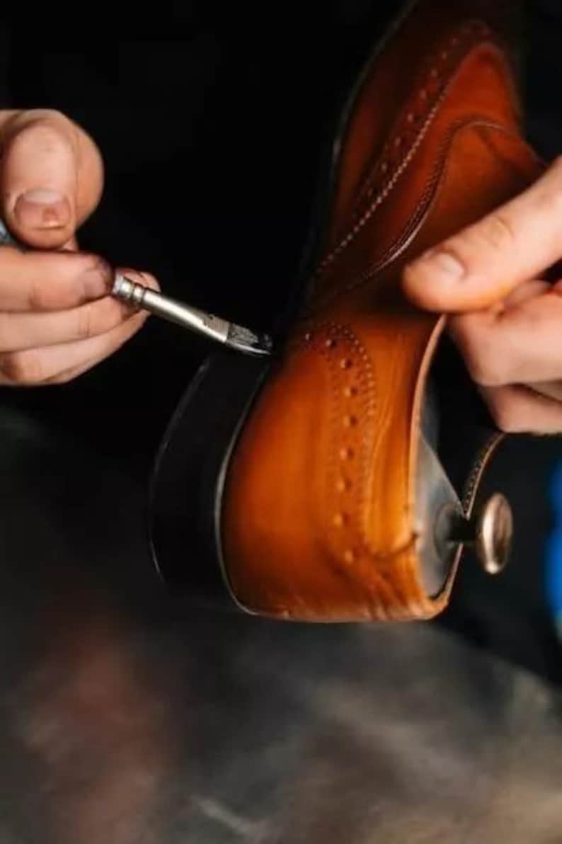 May include: Close-up of a brown leather shoe being worked on with a small brush. The shoe has decorative perforations and a dark sole. The shoemaker is applying a dark substance to the shoe's edge, highlighting the craftsmanship.
