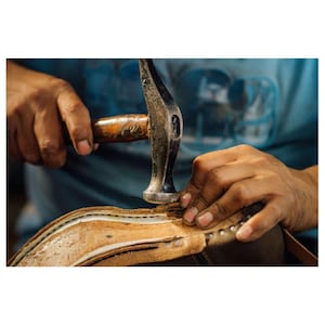 May include: Close-up of a shoemaker hammering a nail into the leather sole of a shoe. The shoemaker's hands are visible, holding the hammer and the shoe. The shoe is made of light brown leather.