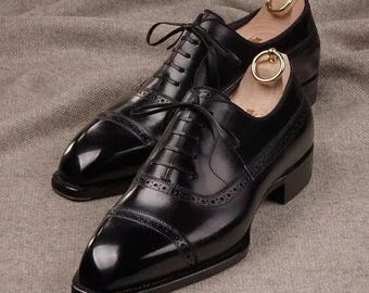 May include: A pair of polished black leather dress shoes with laces, displayed on a neutral-toned surface. The shoes have a classic design with decorative perforations and are paired with wooden shoe trees with gold-colored ring handles.