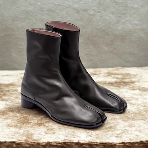 Könnte beinhalten: Ein Paar schwarze Leder-Ankle Boots mit geteiltem Zehendesign. Die Stiefel haben einen niedrigen Blockabsatz und eine glatte, polierte Oberfläche. Das Innenfutter ist hellrosa. Die Stiefel werden auf einer neutralen Oberfläche präsentiert.