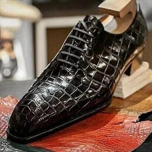 May include: A close-up of a black leather dress shoe with a textured, crocodile-like pattern. The shoe has a pointed toe, laces, and a small heel. The shoe is displayed on a red and brown surface.