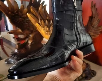 May include: A black ankle boot with a textured, crocodile-like pattern. The boot features a side zipper, a small strap at the top, and a low, square heel. The boot is held by a hand, showcasing its sleek design.