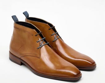 May include: A pair of tan leather chukka boots with blue laces and pull tabs. The boots have a polished finish and a classic design, suitable for various occasions. The soles are a darker brown, providing a contrast to the lighter upper.