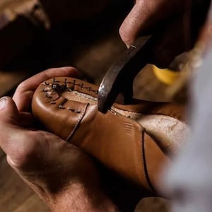 May include: A shoemaker uses a hammer to work on a brown leather shoe. The shoe is held in place by the shoemaker's hands. The stitching is visible on the shoe's upper. The background is a wooden surface.