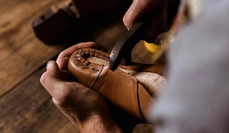 May include: A shoemaker uses a hammer to work on a brown leather shoe. The shoe is held in place by the shoemaker's hands. The stitching is visible on the shoe's upper. The background is a wooden surface.