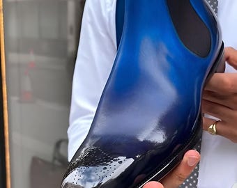 May include: A blue and black leather Chelsea boot with a glossy finish. The boot features a pointed toe with decorative perforations and elastic side panels. The upper transitions from a deep blue to black, creating a gradient effect. The sole is black.
