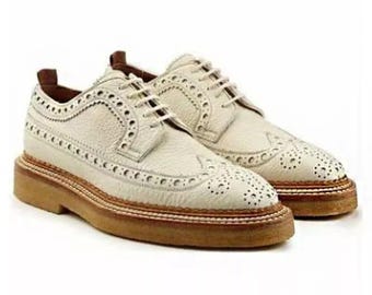May include: A pair of cream-colored brogue shoes with decorative perforations and white laces. The shoes have a thick, light brown sole with a stitched border. The uppers are made of textured leather.