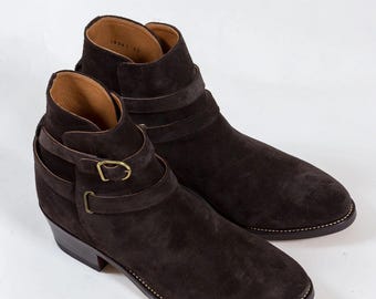 May include: A pair of dark brown suede ankle boots. The boots feature a strap and buckle detail, a low heel, and a stitched sole. The interior lining is a lighter brown color. The boots are displayed on a white background.