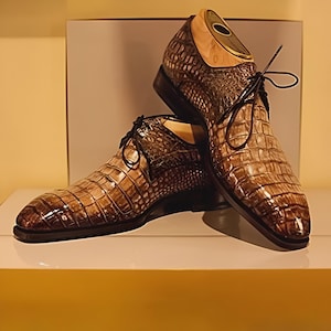 May include: A pair of brown leather dress shoes with a crocodile skin texture. The shoes have black laces and soles, and are displayed with wooden shoe trees inside. The shoes are on a white surface.