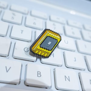 Può includere: Una spilla rettangolare in smalto con un design giallo e nero, con un centro grigio con un punto esclamativo, poggia su una tastiera bianca di un laptop. La spilla ha un'estetica pixelata.