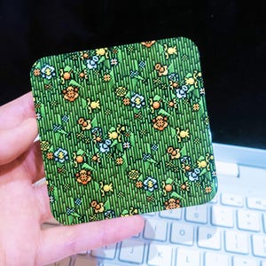 Könnte beinhalten: Ein quadratischer Untersetzer mit einem pixeligen Design, das ein grünes Grasfeld mit orangefarbenen, gelben und blauen Blumen zeigt. Der Untersetzer wird in einer Hand gehalten, mit einem weißen Laptop im Hintergrund.