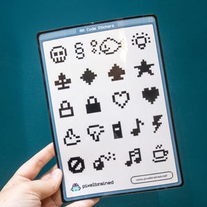 以下が含まれることがあります： ドット絵のコードステッカーシート。ドクロ、音符、コーヒーカップなど、様々な記号が描かれています。ステッカーは白黒で、「Att Code Stickers」の文字が上部にあります。「pixelbrained.net」のウェブサイトが下部に印刷されています。