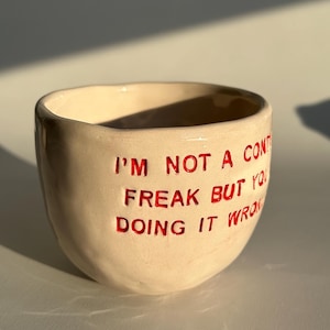Op de afbeelding: Een beige keramische mok met een onregelmatige, handgevormde vorm. De buitenkant is voorzien van rode tekst in reliëf die luidt: I'M NOT A CONTROL FREAK BUT YOU'RE DOING IT WRONG. Het oppervlak heeft een gladde, glanzende afwerking met subtiele deukjes.
