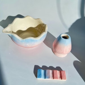 May include: A ceramic matcha bowl with a wavy rim, a matching whisk holder, and a rectangular rest, all featuring a gradient of soft blue and pink hues. The interior of the bowl displays embossed text, and the surfaces have a glossy finish.
