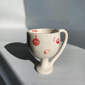 Może przedstawiać: Biały ceramiczny kubek z podstawą na cokole i zakrzywionym uchwytem. Kubek jest ozdobiony czerwonymi ilustracjami kuli dyskotekowej, koktajlu i oliwki. Numer "27.624" jest wydrukowany na czerwono w pobliżu krawędzi.